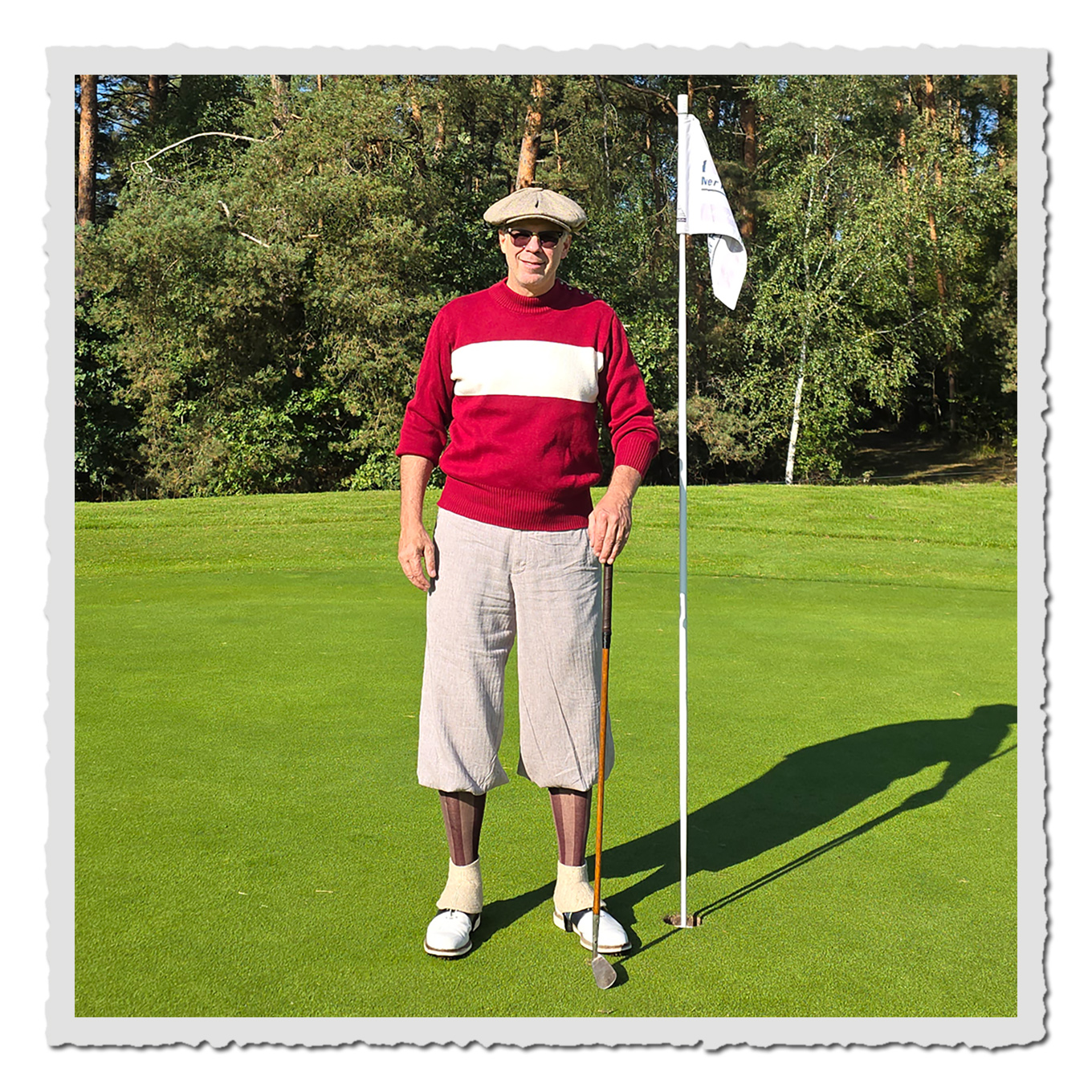 Mann in graubrauner Knickerbocker, rotem Pullover und Schiebermütze auf grünem Golfplatz neben Fahne.